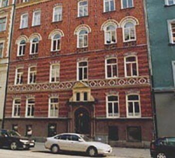 Rött tegelhus i fyra våningar med ljus stenornamentik kring rundbågade fönster och en valvd entréportal, två bilar parkerade vid trottoarkanten.