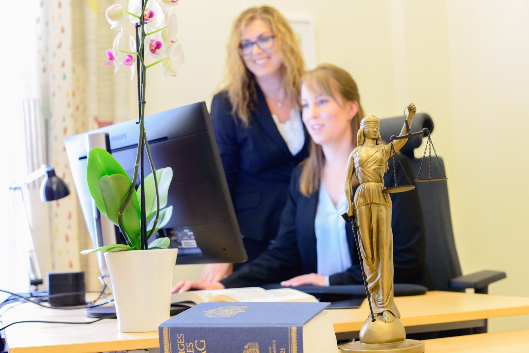 Två medarbetare vid en skrivbordsdator på byrån. På bordet står en mässingsfigur av Justitia och en orkidé.